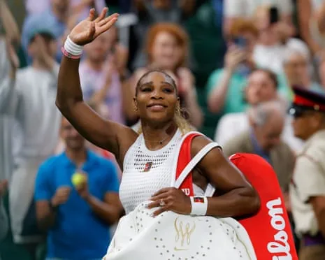 Serena Williams in white outfit waves to the crowd while holding a red Wilson bag