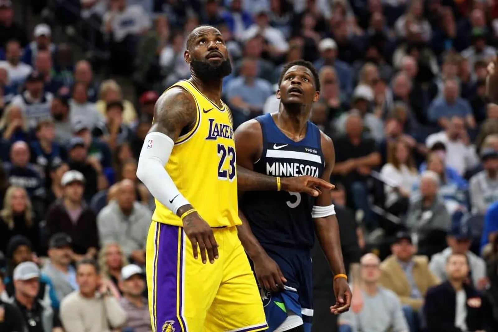 LeBron James of the Los Angeles Lakers, wearing No. 23 on his jersey, holds his arm out in front of Anthony Edwards of the Minnesota Timberwolves while both look up with the crowd in the background.