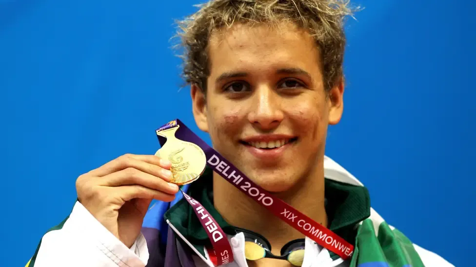 A head and shoulders shot of Chad le Clos shows him smiling as he holds up a gold medal on a dark red Delhi 2010 ribbon with his right hand. He wears a predominantly white and green tracksuit jacket and a pair of swimming goggles are visible around his neck