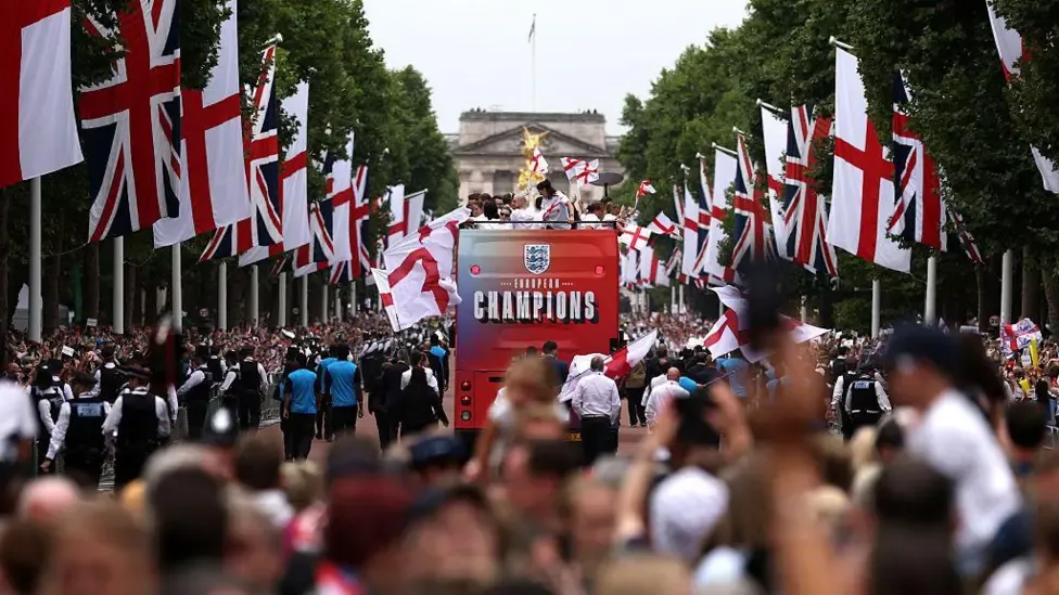 An open-top England women's victory parade bus, displaying "EUROPEAN CHAMPIONS" on the back, driving down The Mall in London. The tree-lined street is heavily decorated with Union Jacks and St George's Cross flags, with a large crowd watching and Buckingham Palace visible in the background.